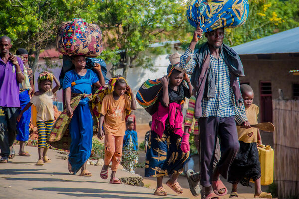 People walk along a dirt path carrying large bundles wrapped in colorful fabric and a yellow jerrycan, with trees and houses in the background.